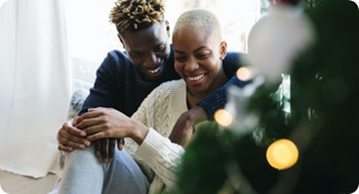 Couple embracing and smiling, seated on the floor next to a Christmas tree.